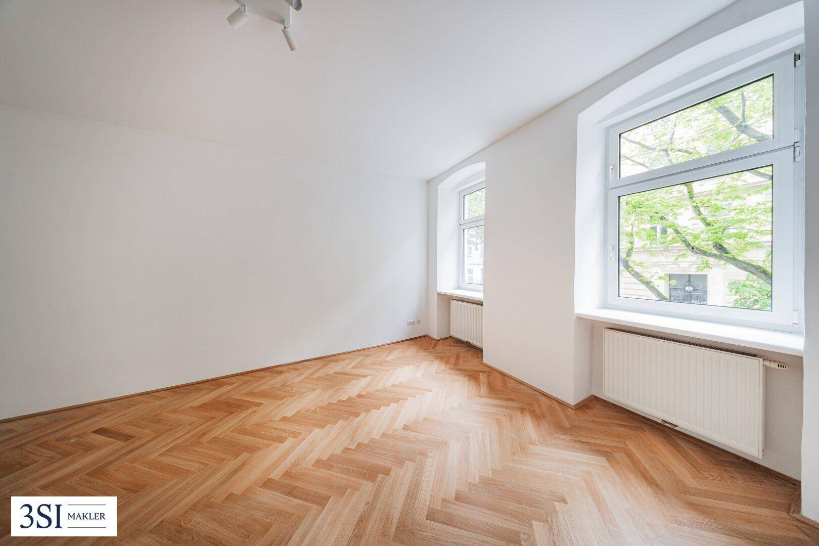 Geräumiges Zimmer mit hellem Fischgrätparkett und großen Fenstern, die einen Blick ins Grüne bieten.