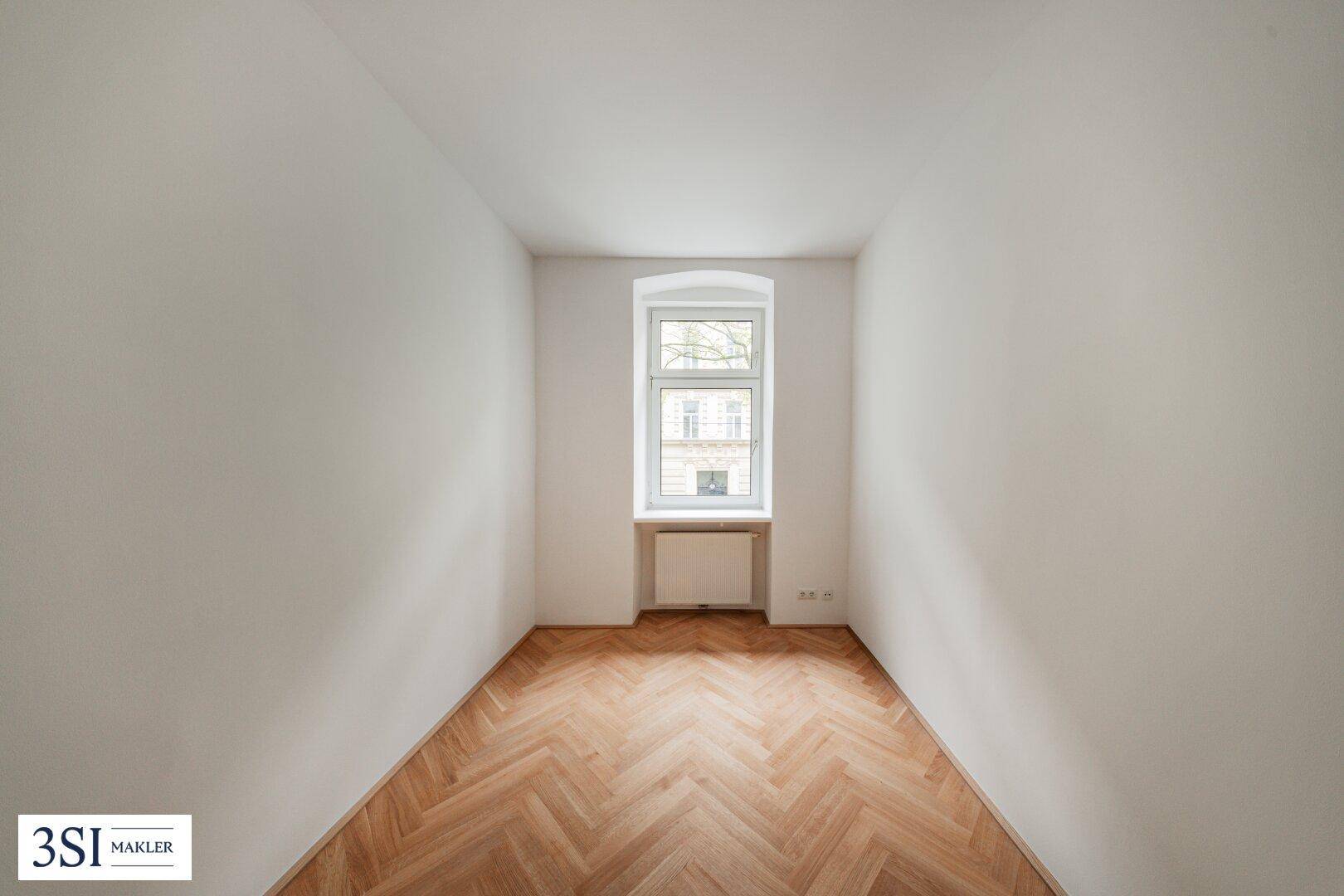 Helles, leeres Zimmer mit Fischgrätparkettboden und einem Fenster mit Blick nach draußen.