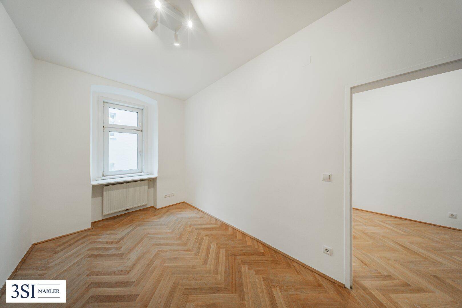 Helles Zimmer mit Fischgrätparkett und einem Fenster, das den Raum mit natürlichem Licht versorgt.
