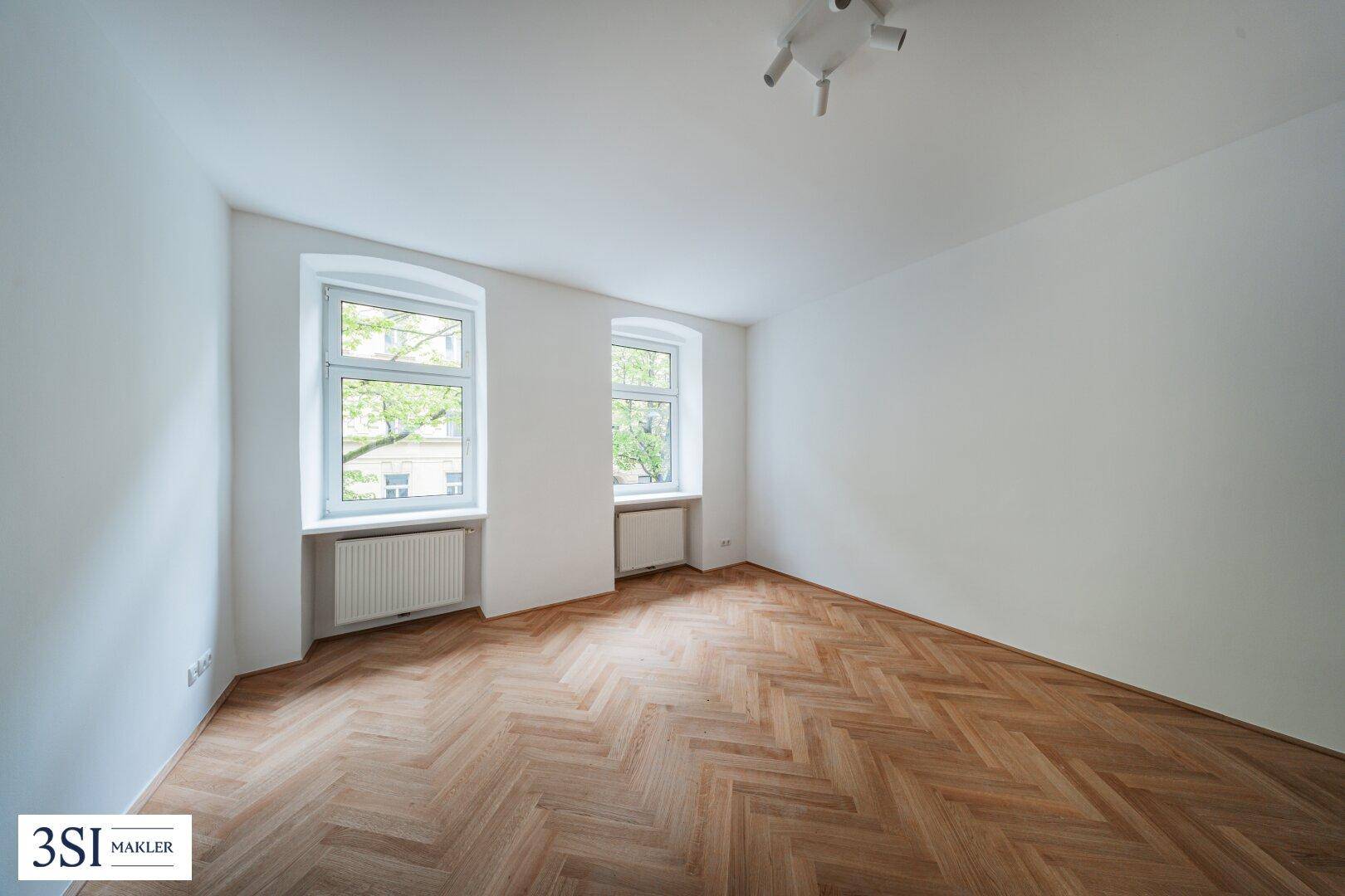 Geräumiges, helles Zimmer mit Fischgrätparkett und zwei Fenstern, die einen Blick ins Grüne ermöglichen.