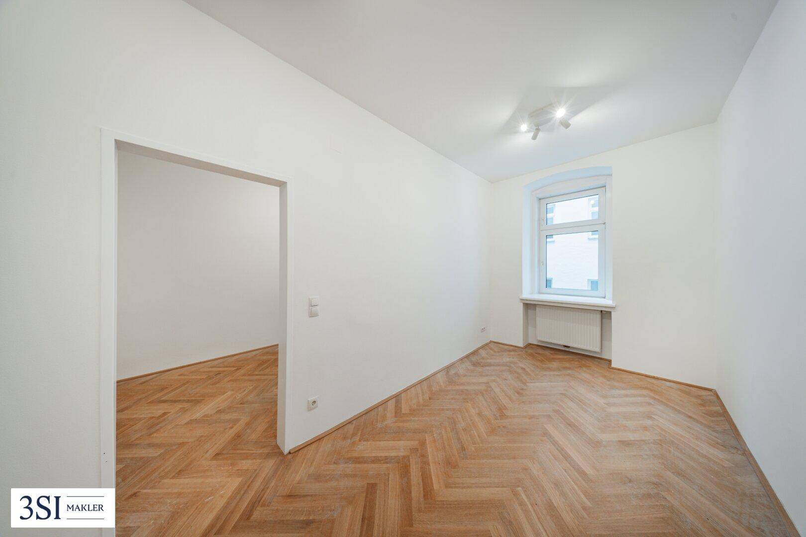 Leeres Zimmer mit Fischgrätparkett und einem Fenster, das den Raum mit Tageslicht erfüllt.