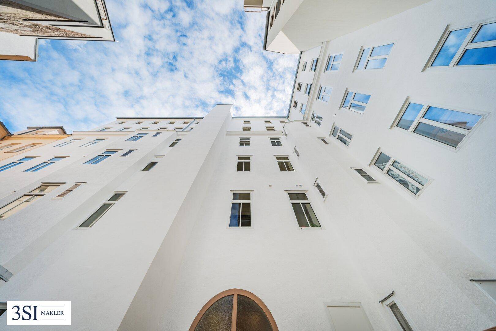 Blick nach oben auf die weiße Fassade eines Gebäudes in einem Innenhof unter blauem Himmel.