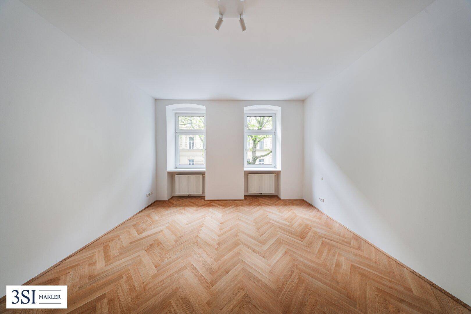 Leeres Schlafzimmer mit klassischem Fischgrätparkett und zwei großen Fenstern, die viel Licht hereinlassen.