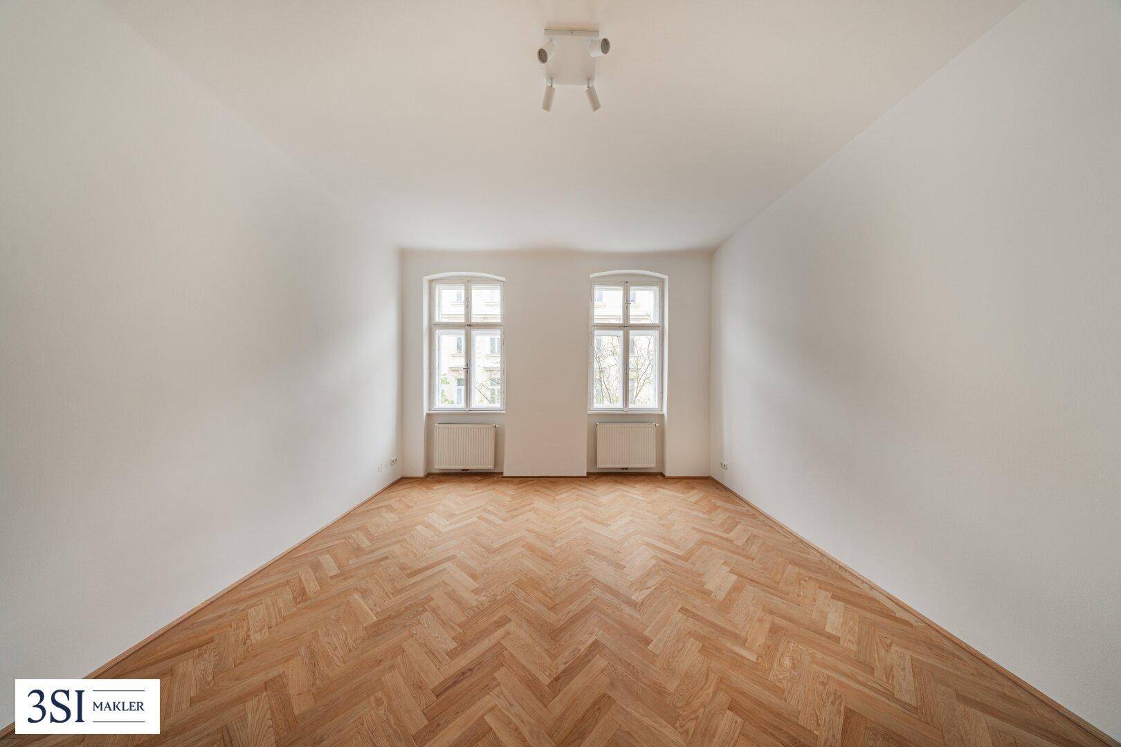 Geräumiger, heller Raum mit Fischgrätparkett und zwei großen Fenstern, die viel natürliches Licht hereinlassen.