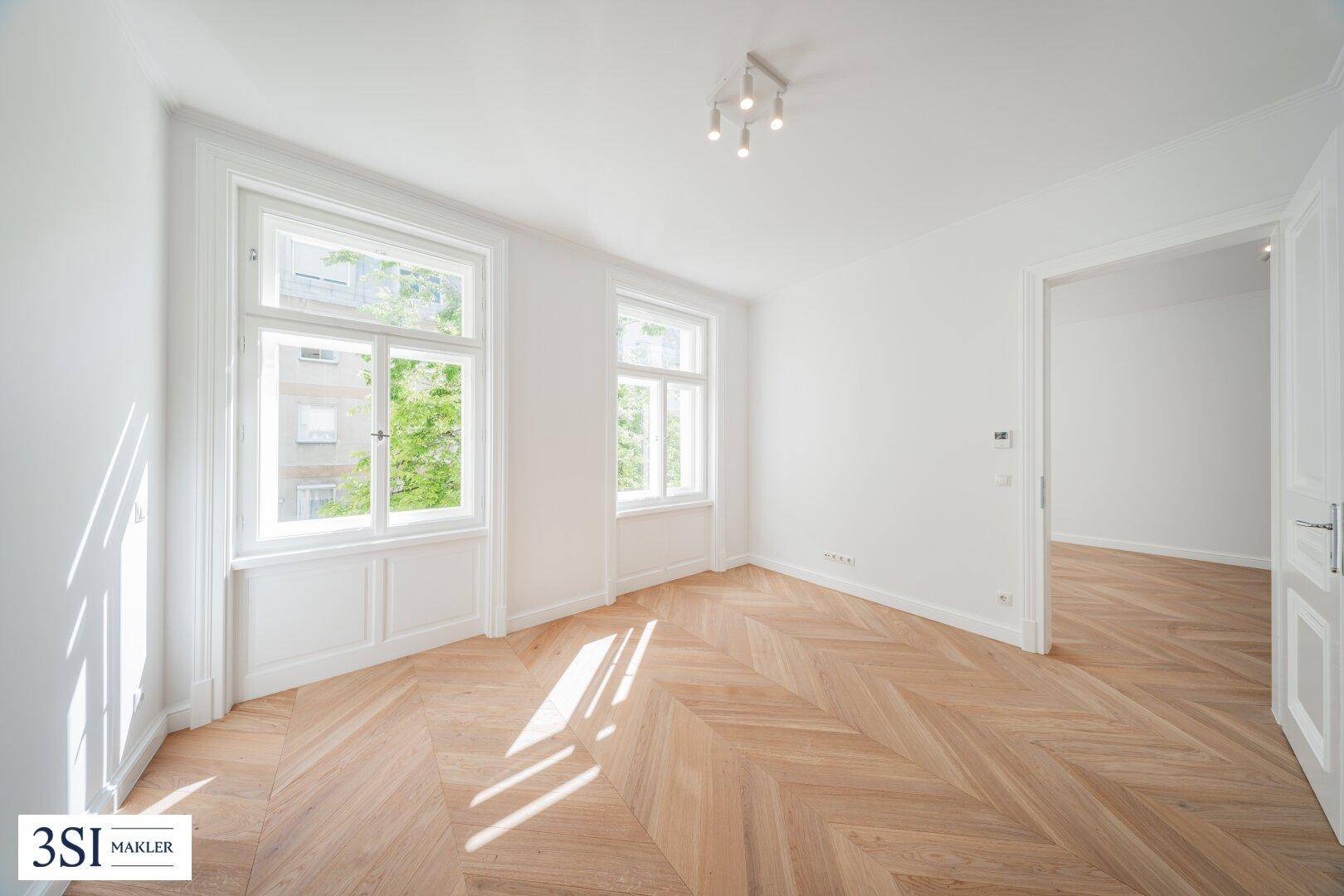 Heller Raum mit Fischgrätparkettboden, großen Fenstern und viel natürlichem Tageslicht.