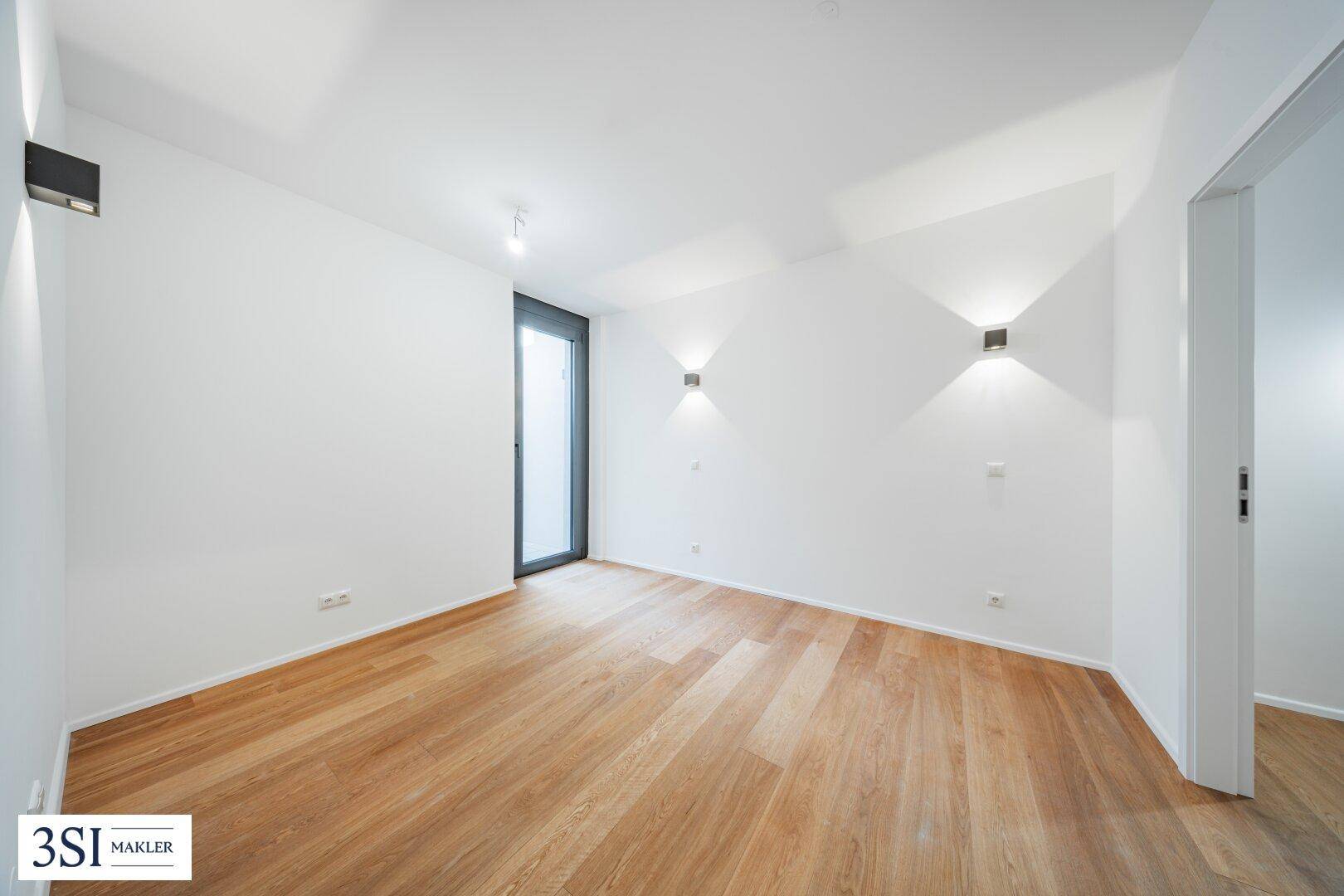 Leeres Zimmer mit Holzboden, weißen Wänden und einer hohen Tür, die nach draußen führt.
