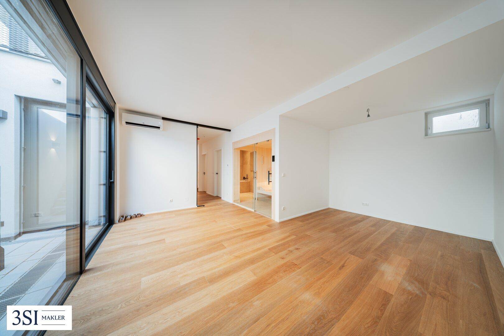 Geräumiger Raum mit Holzboden, großen Fenstern und einer Glastür, die zu einem Badezimmer führt.