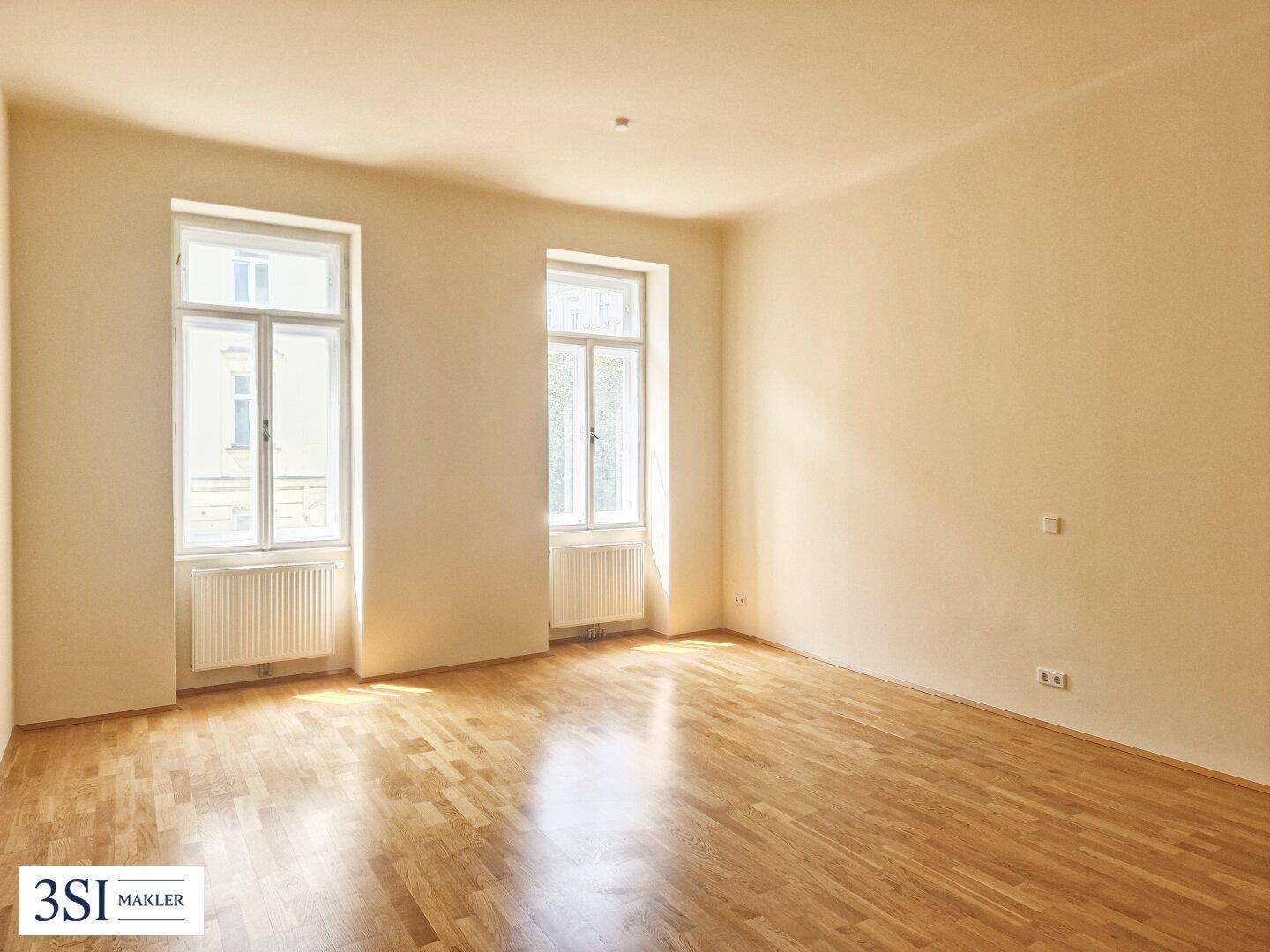 Geräumiges Zimmer mit Holzboden und viel Tageslicht durch zwei hohe Fenster, ideal für Wohnzwecke.