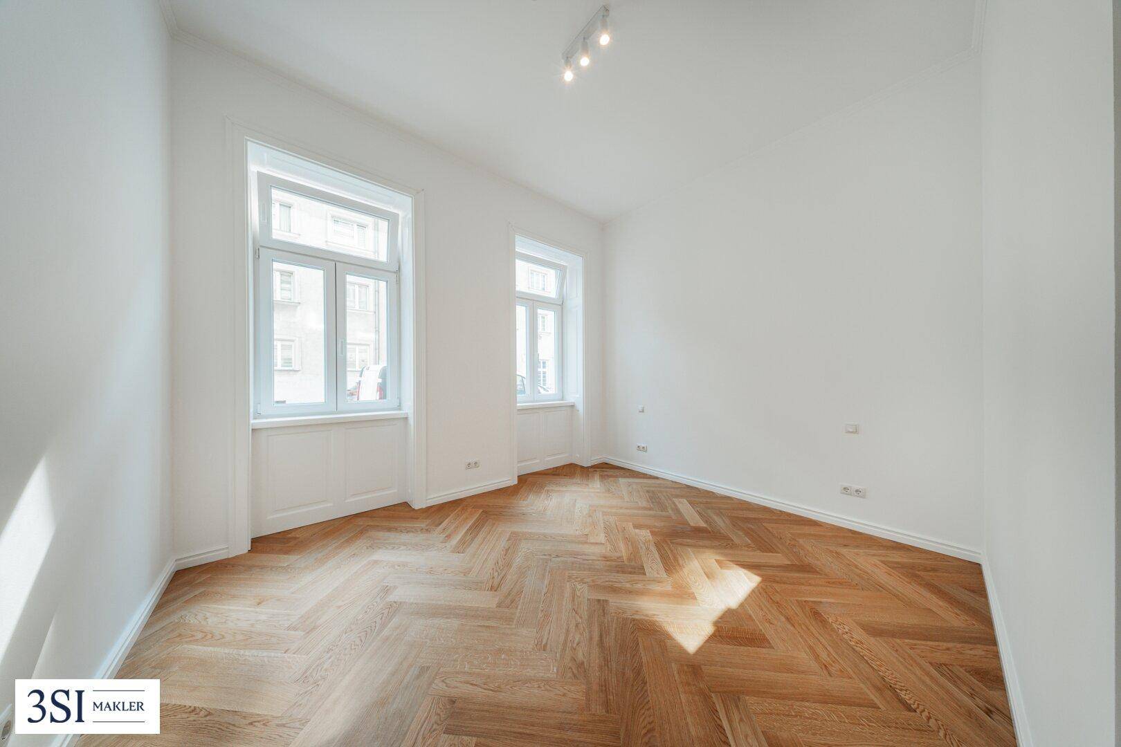 Lichtdurchfluteter Raum mit klassischem Fischgrätparkett und hohen Fenstern, ideal für ein Schlaf- oder Arbeitszimmer.