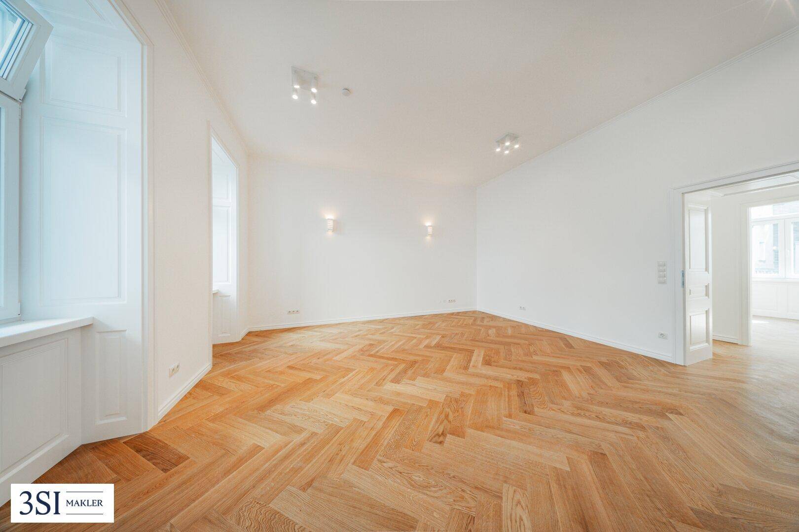 Großzügiges Zimmer mit Fischgrätparkett und viel Tageslicht durch hohe Fenster.