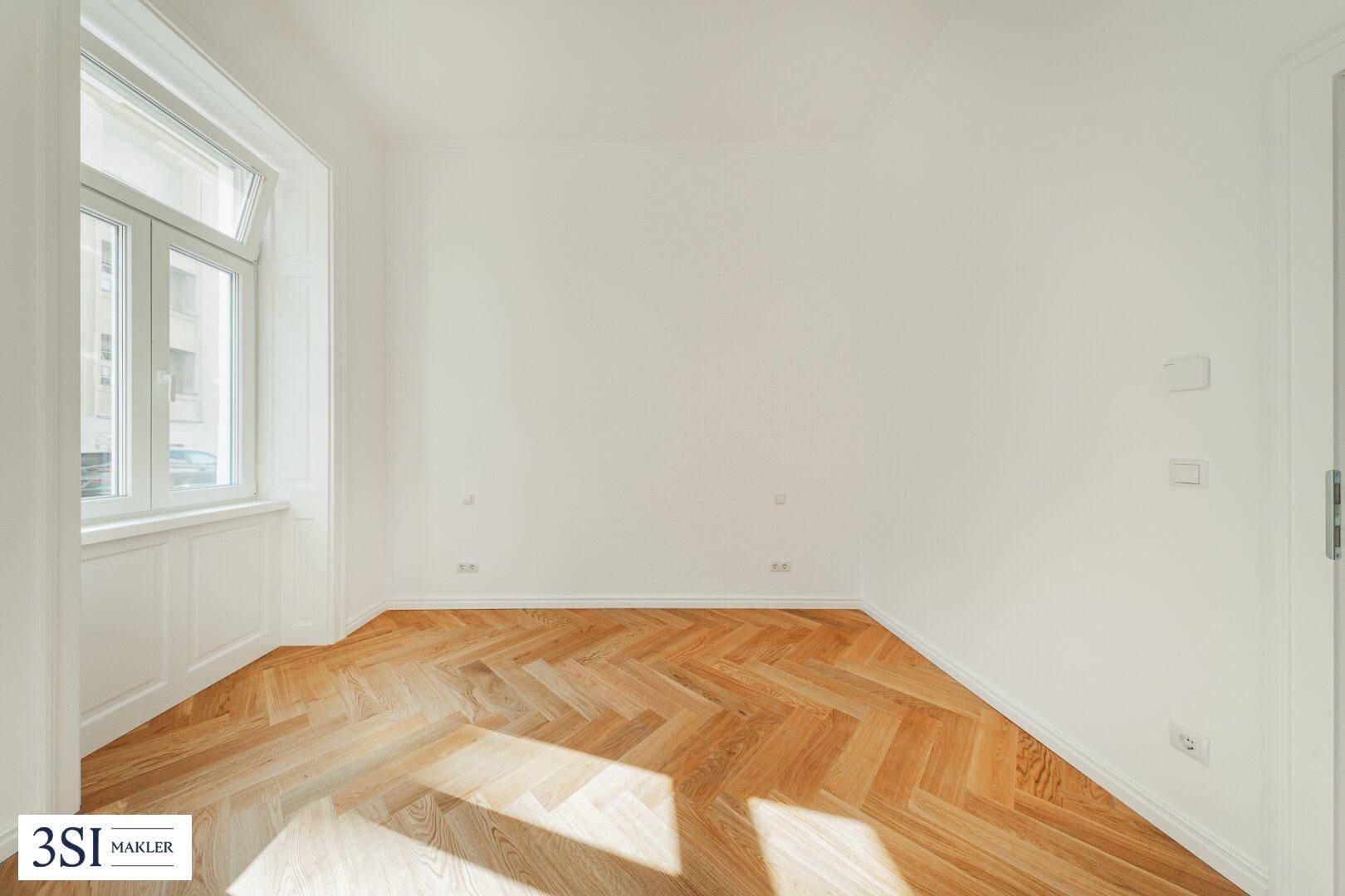 Helles Zimmer mit Fischgrätparkett und einem Fenster, das von Sonnenlicht durchflutet wird.