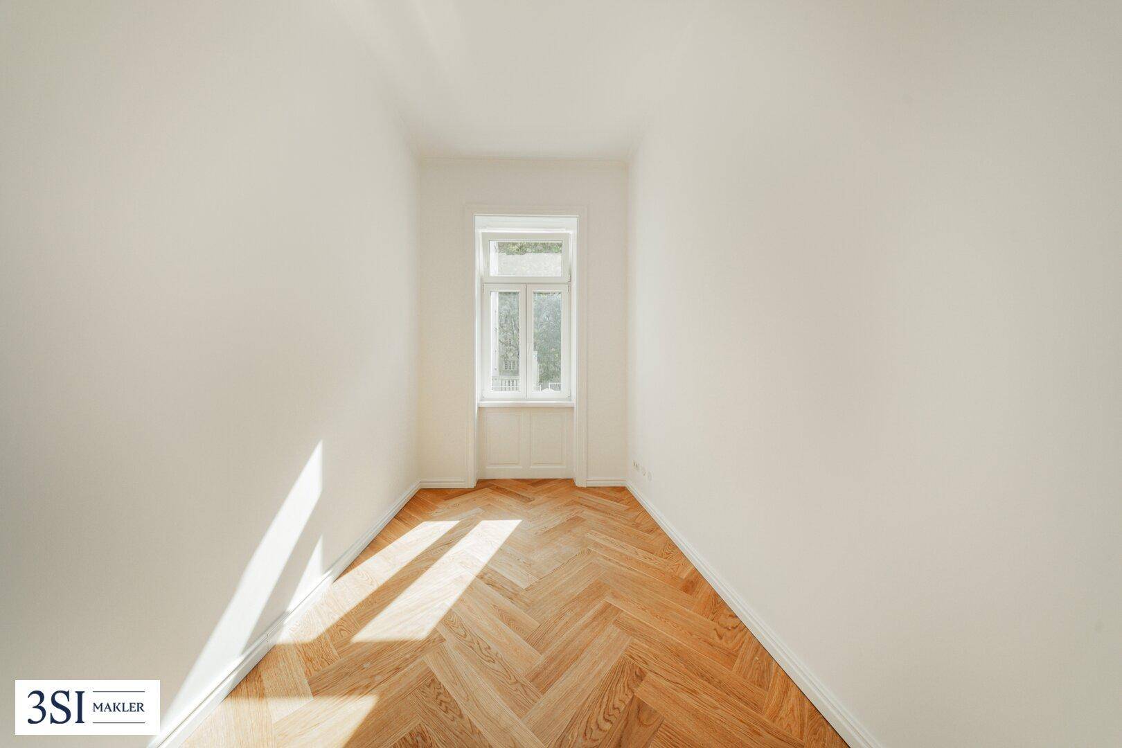 Ruhiges Zimmer mit Fischgrätparkett und einem Fenster, das von der Sonne beschienen wird.