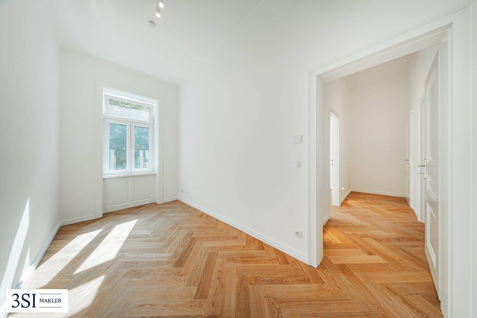 Geräumiges Zimmer mit Fischgrätparkett, einem Fenster und offenem Zugang zu einem weiteren Bereich.