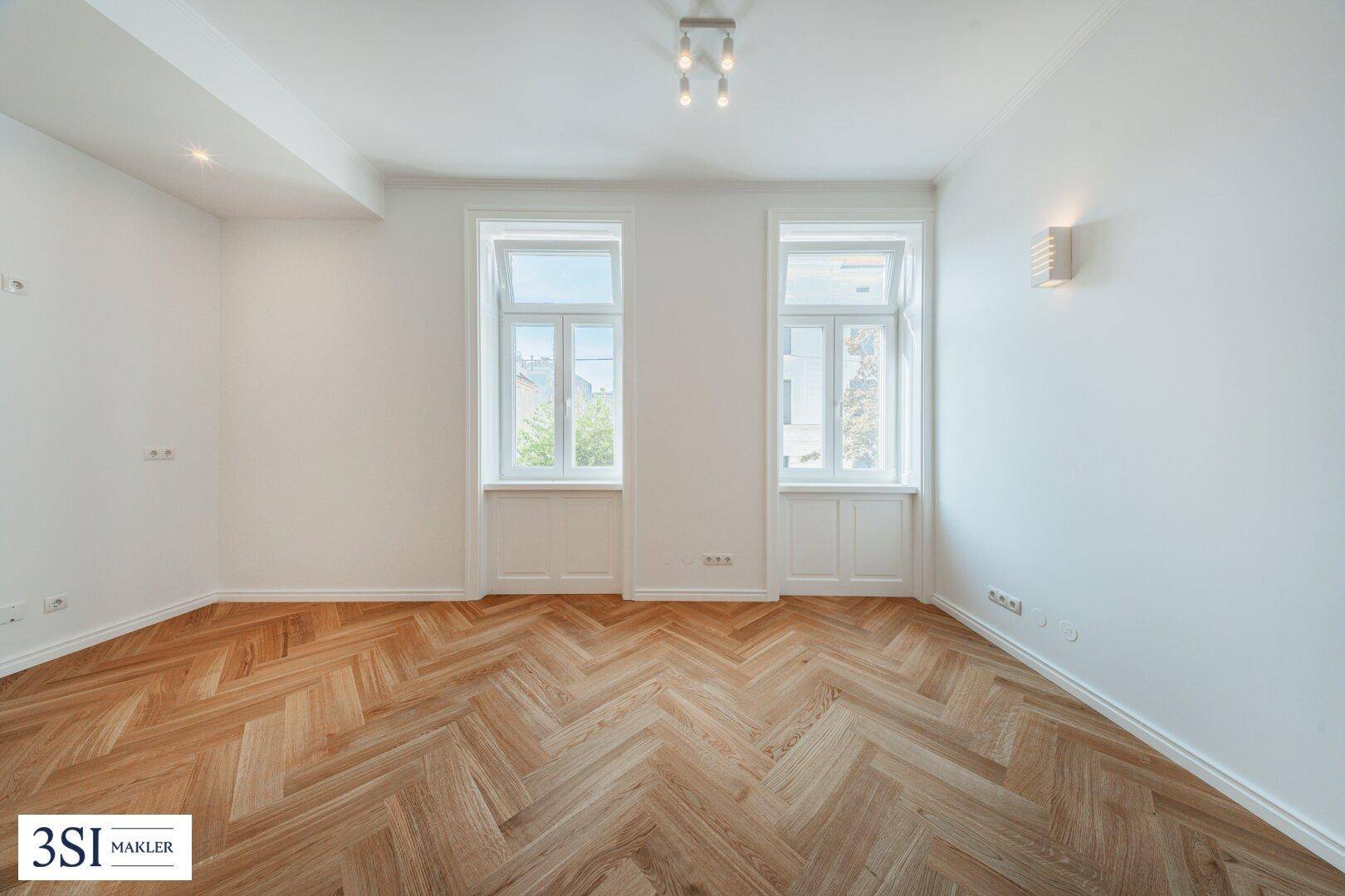 Leerer, heller Raum mit Fischgrätparkett und zwei hohen Fenstern, bereit für individuelle Einrichtung.