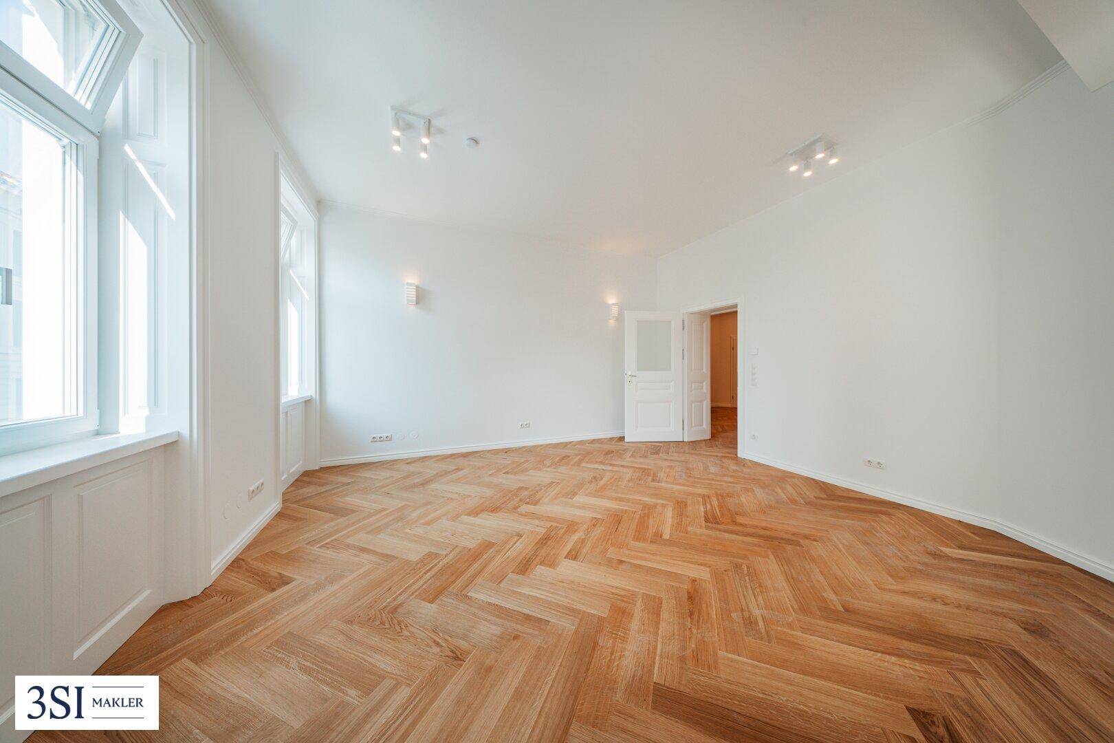 Geräumiges Wohnzimmer mit elegantem Fischgrätparkett und viel Tageslicht durch große Fenster.