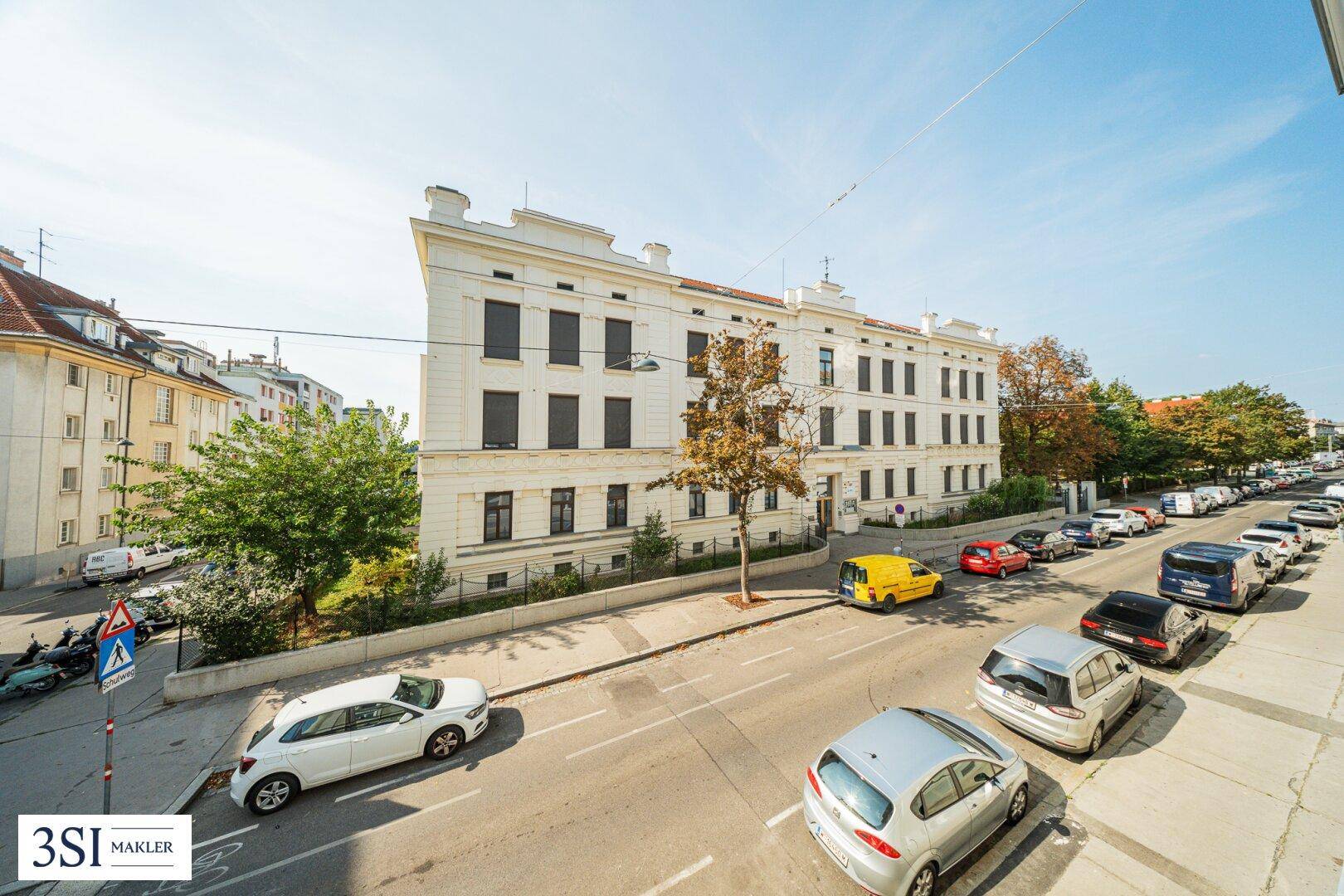 Blick auf die Straße mit parkenden Autos und einem klassischen Wohngebäude unter blauem Himmel.