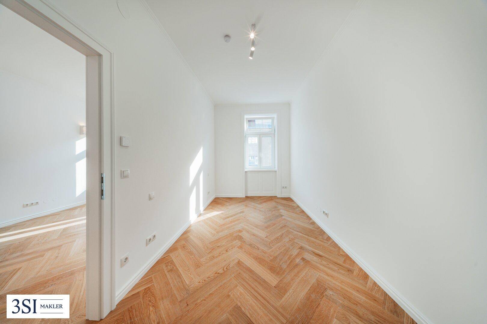 Langes, helles Zimmer mit Fischgrätparkett und einem Fenster, das viel Sonnenlicht hereinlässt.