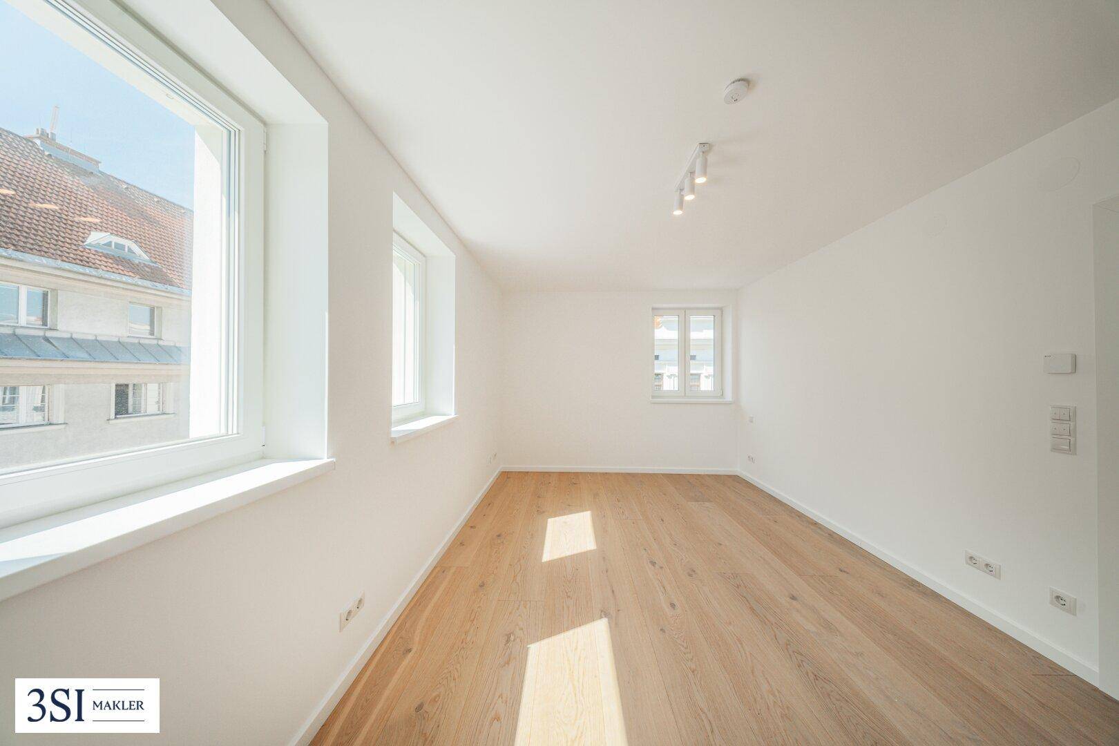 Heller Raum mit Holzboden und großen Fenstern, die viel Sonnenlicht hereinlassen.