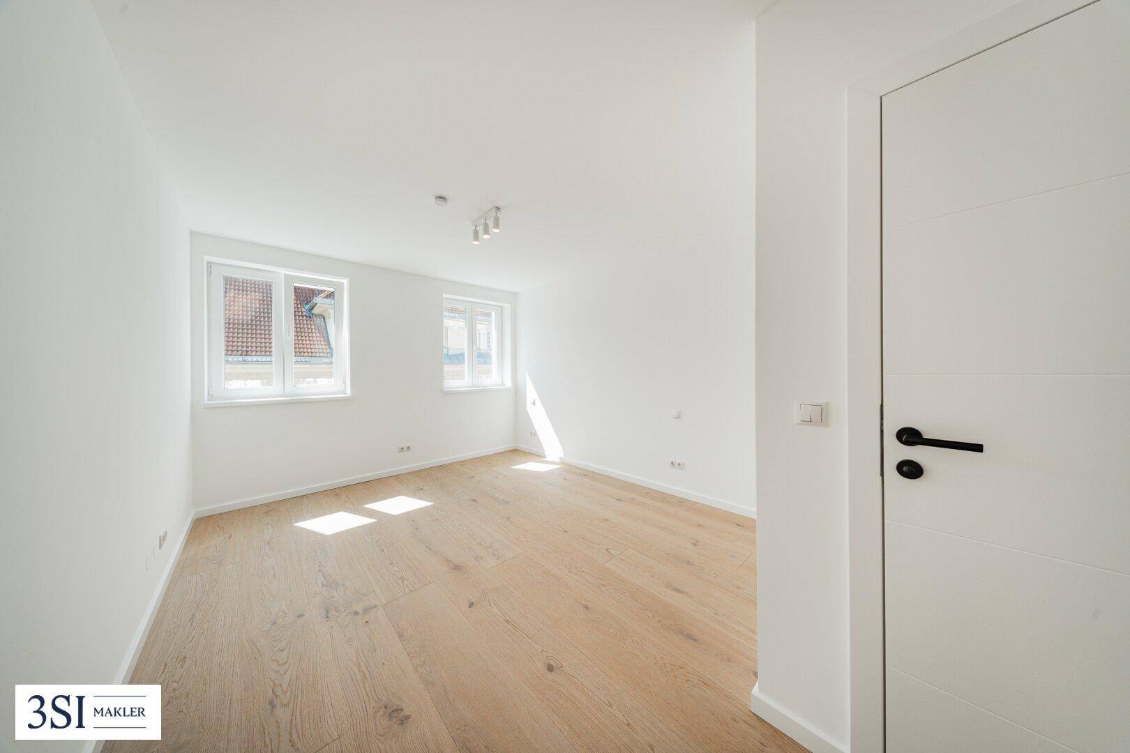 Freundliches, leeres Schlafzimmer mit Holzboden und zwei Fenstern, die Sonnenlicht hereinlassen.