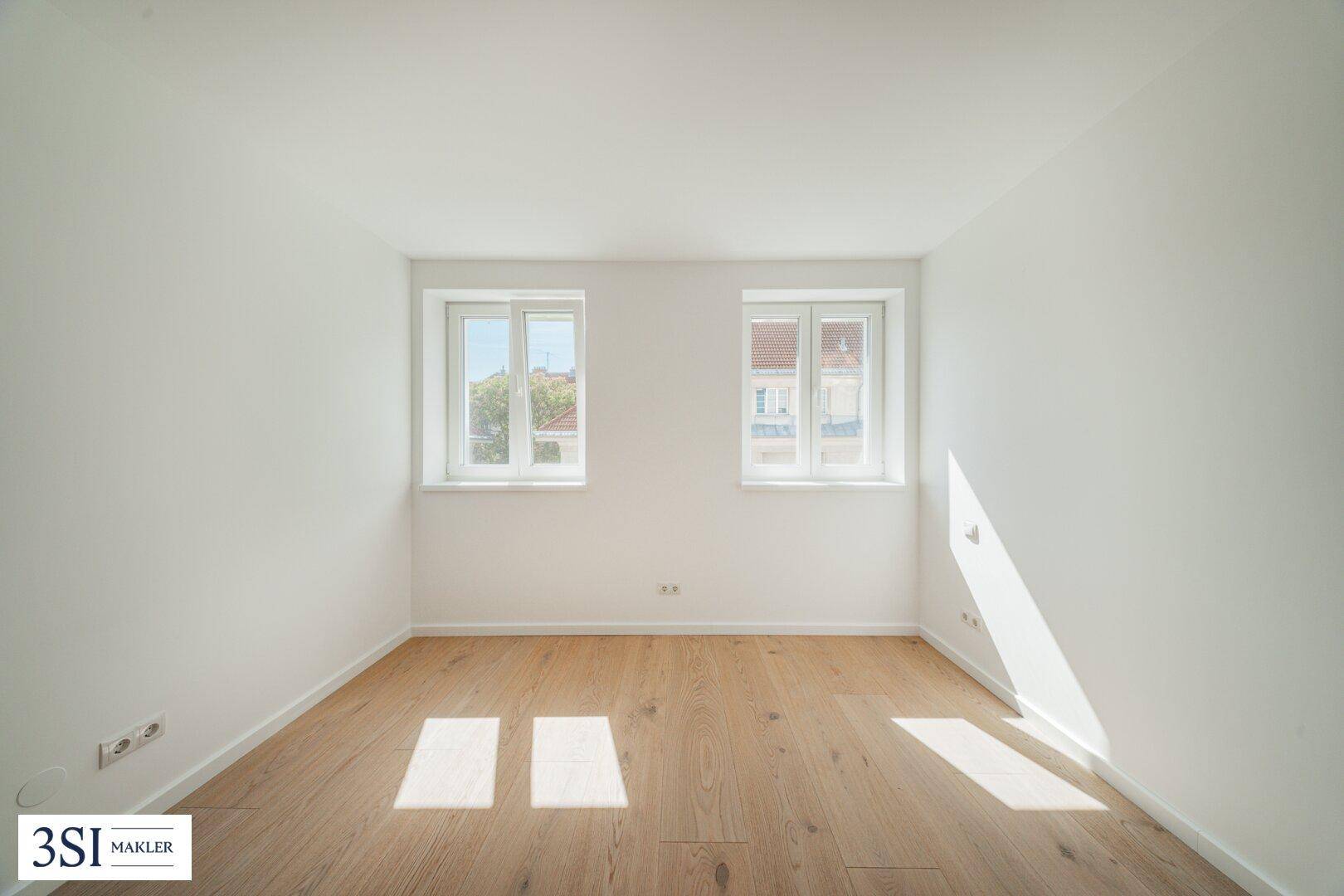 Helles, leeres Zimmer mit Holzboden und zwei Fenstern, die viel Tageslicht hereinlassen.
