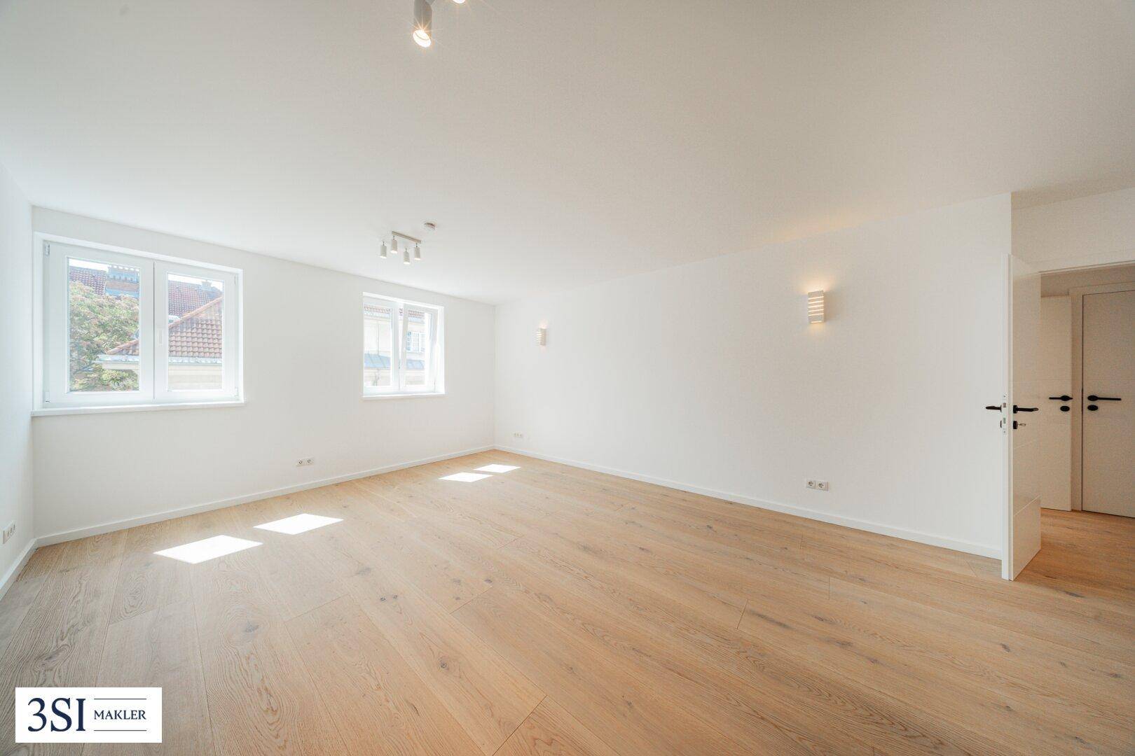 Helles, leeres Zimmer mit Holzboden und großen Fenstern, das viel Tageslicht hereinlässt.