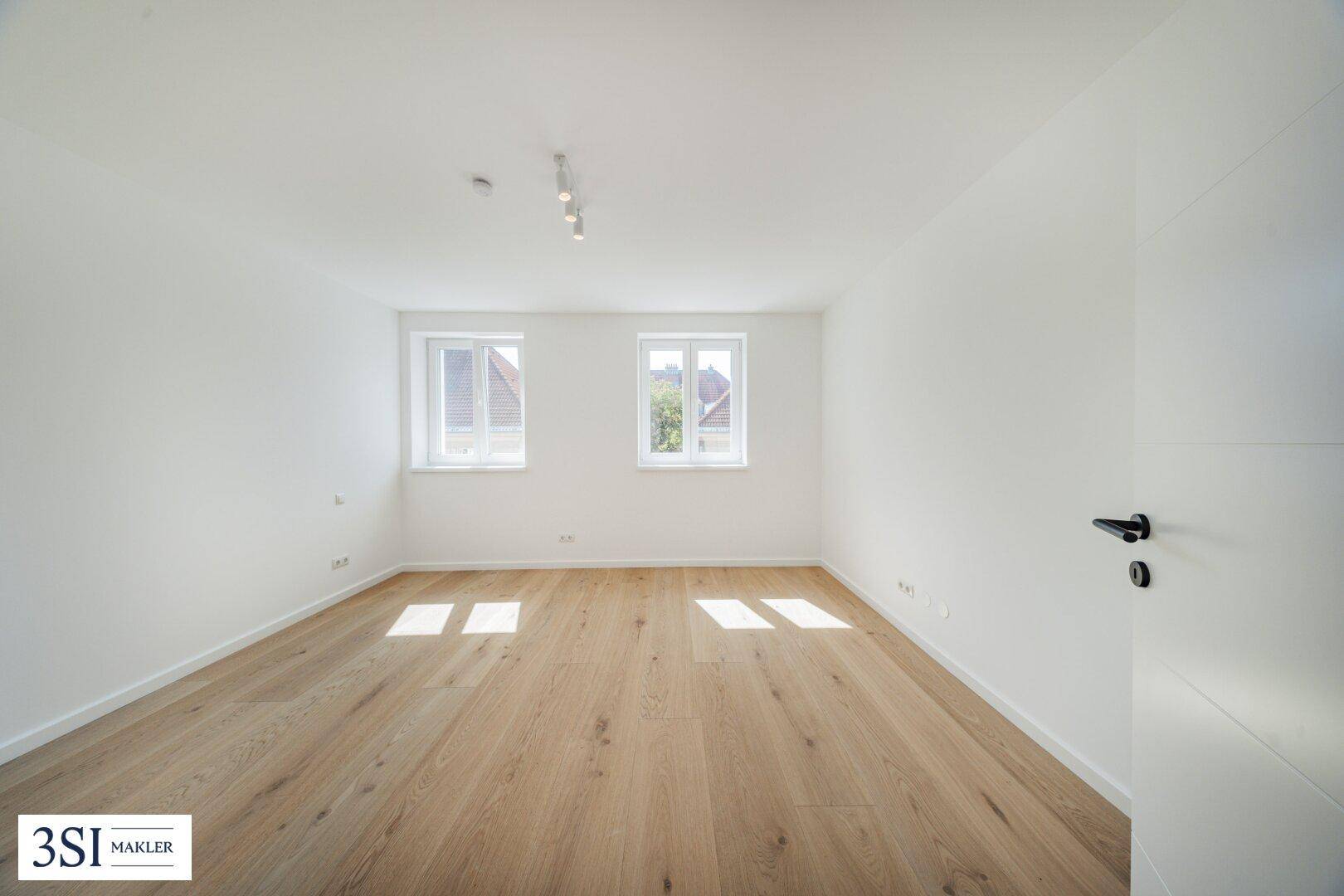 Leerer, lichtdurchfluteter Raum mit hellem Holzboden und zwei großen Fenstern, ideal für individuelle Gestaltung.