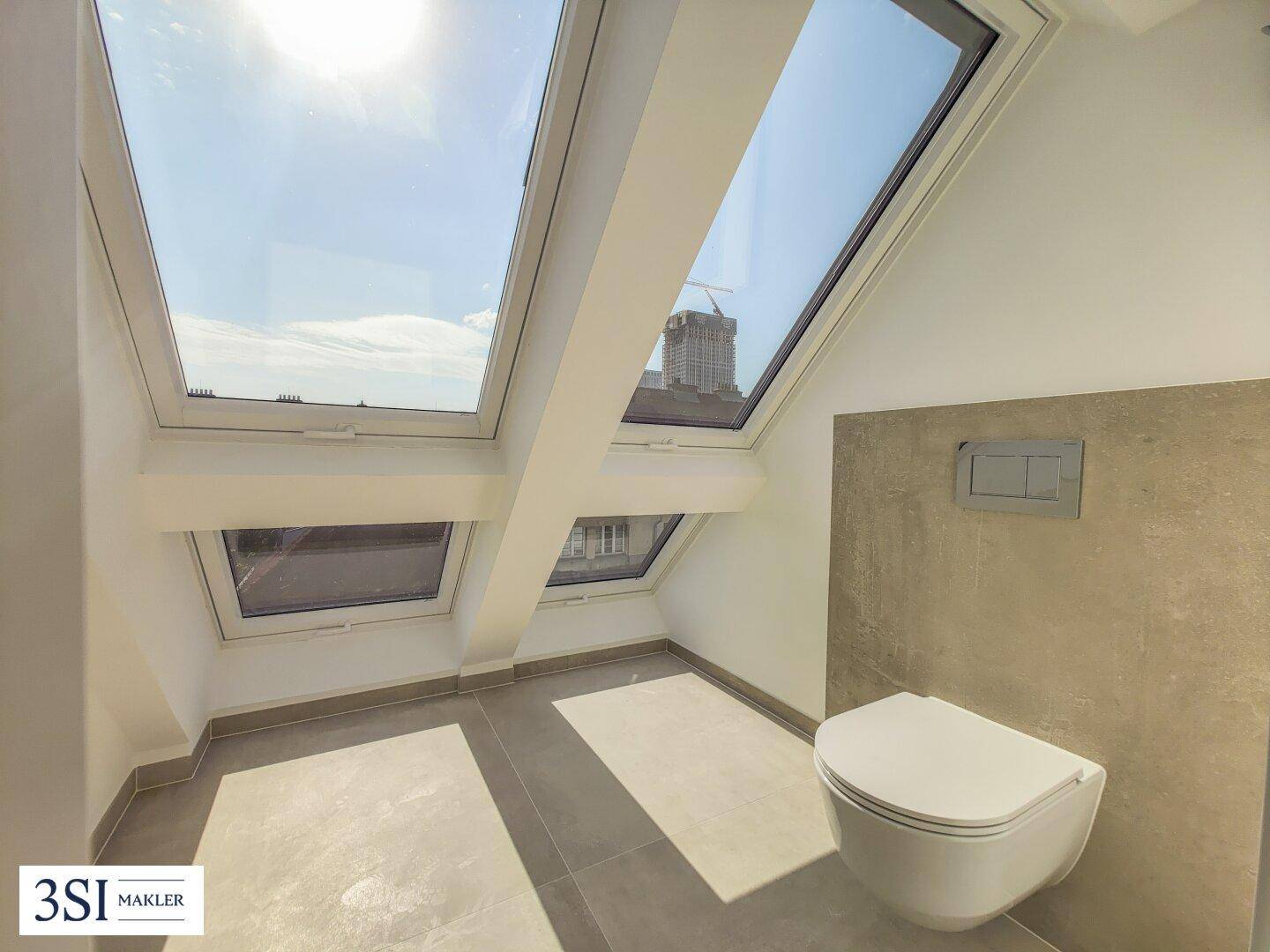 Modernes WC im Dachgeschoss mit Schrägfenstern, grauen Fliesen und wandhängender Toilette.
