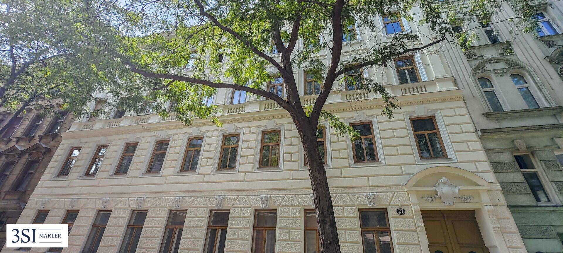 Klassische Fassade eines Altbaus mit hohen Bäumen, die Schatten spenden und die Straße säumen.