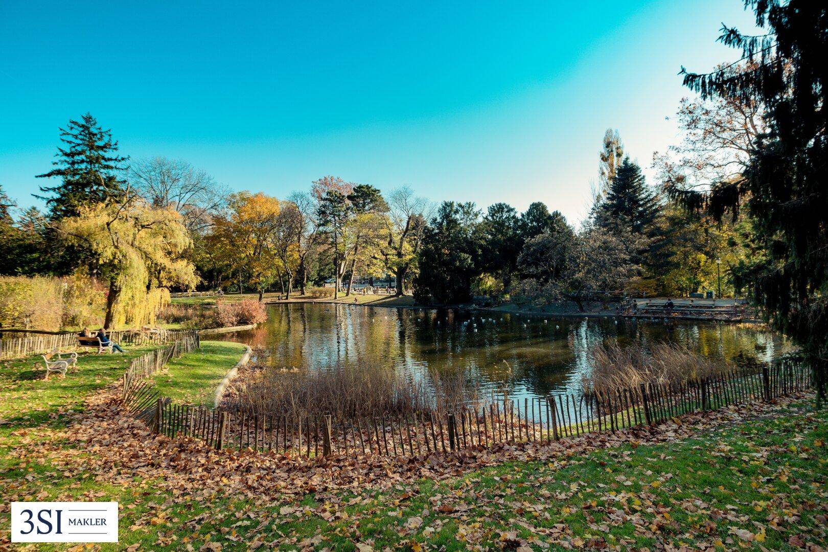 Idyllischer Park mit einem Teich, umgeben von Bäumen und herbstlichem Laub unter blauem Himmel.