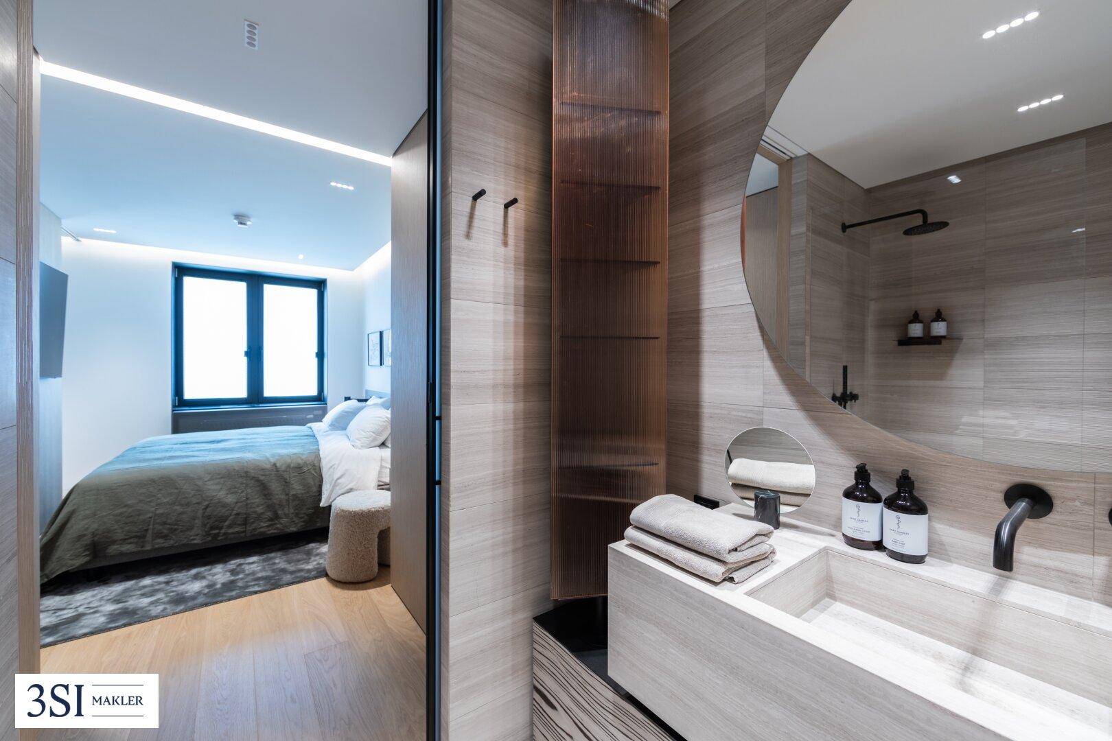 Modernes Badezimmer mit eleganten Fliesen, rundem Spiegel und direktem Zugang zum Schlafzimmer.