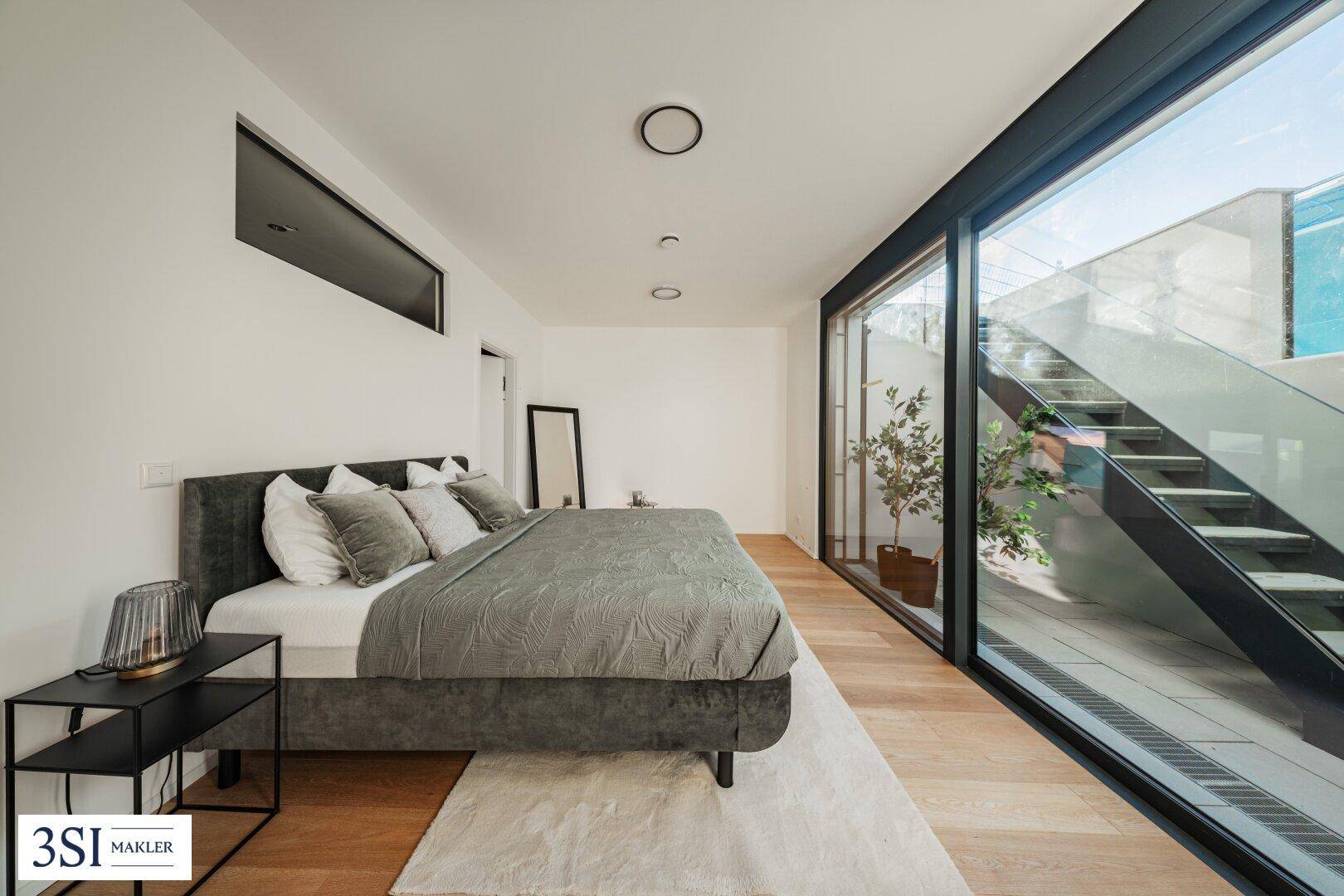 Modernes Schlafzimmer mit großem Bett, Holzboden und raumhohen Fenstern mit Blick auf den Außenbereich.
