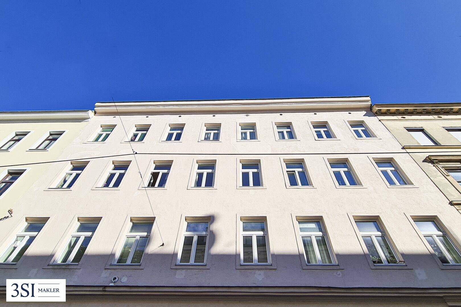 Ansicht der hellen Fassade eines Mehrfamilienhauses mit vielen Fenstern unter blauem Himmel.