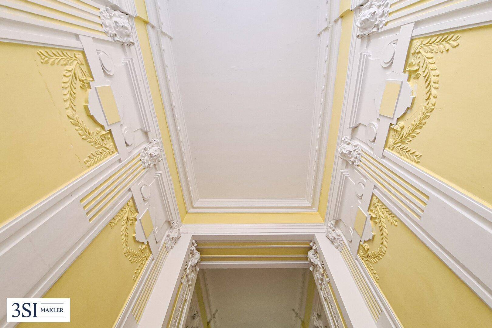 Historisches Treppenhaus mit aufwendigen Stuckverzierungen und gelben Wänden.