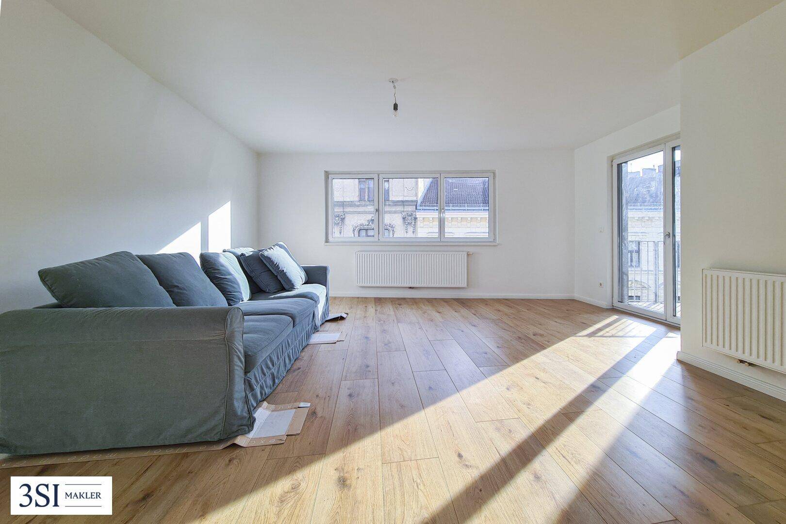Helles Wohnzimmer mit Holzboden, einem Sofa und Zugang zu einem Balkon, durchflutet von Sonnenlicht.