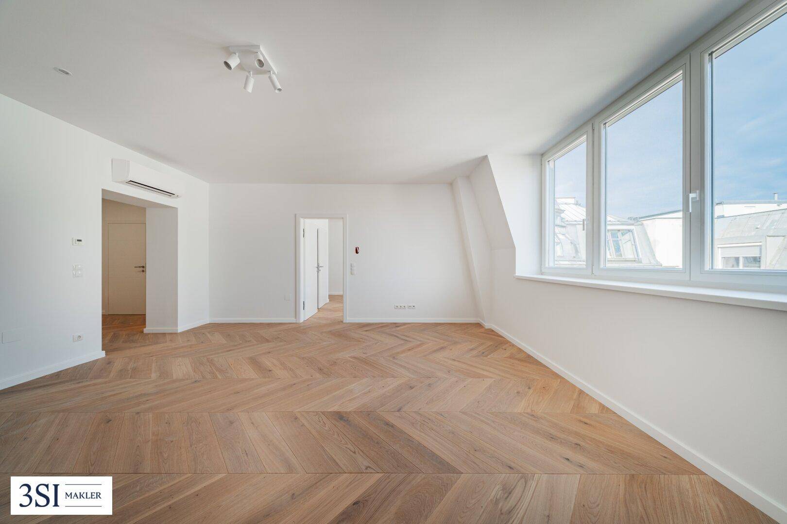 Geräumiger, heller Raum mit elegantem Fischgrätparkett und großen Fenstern, die viel Tageslicht hereinlassen.