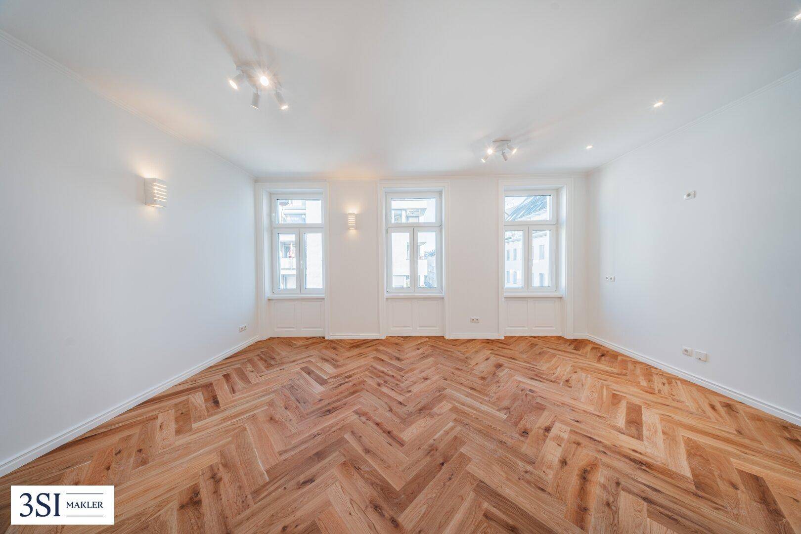 Geräumiges, lichtdurchflutetes Zimmer mit elegantem Fischgrätparkett und weiß gestrichenen Wänden.