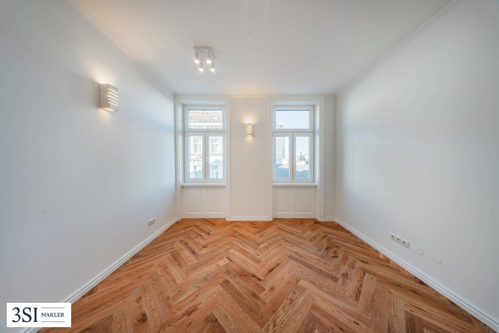 Heller Raum mit elegantem Fischgrätparkett und zwei großen Fenstern, frisch renoviert.