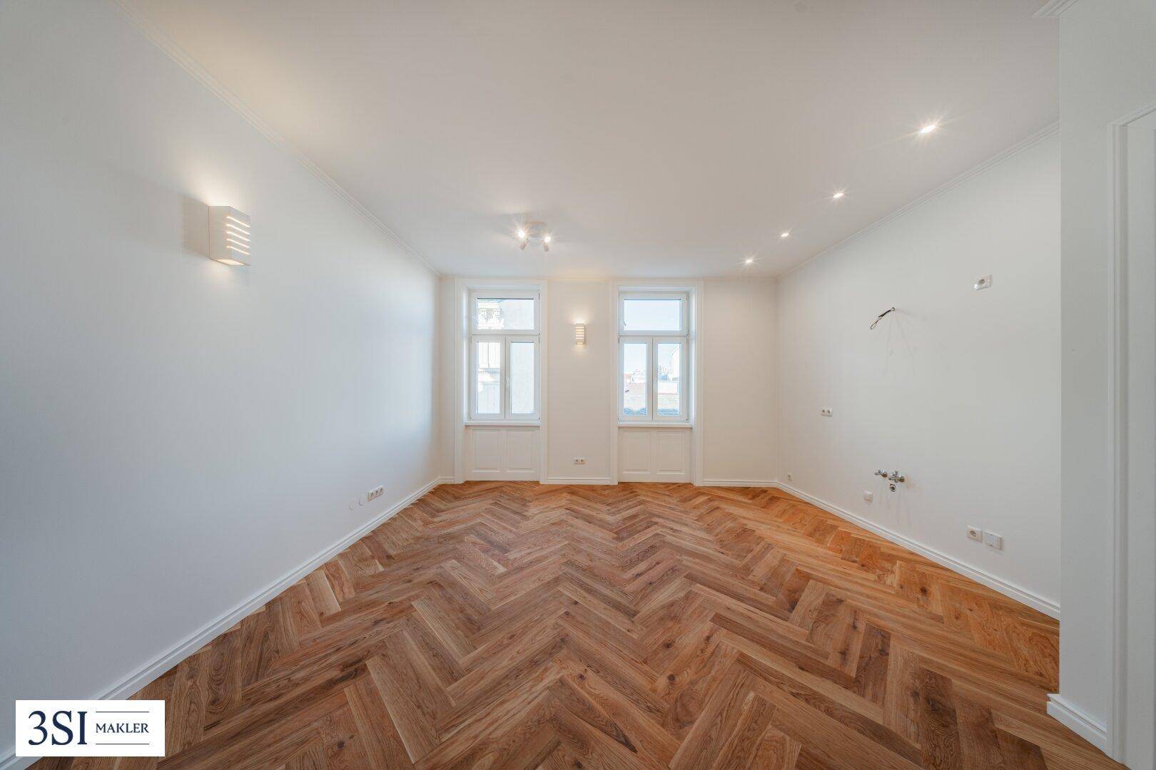 Geräumiges, lichtdurchflutetes Zimmer mit elegantem Fischgrätparkett und weiß gestrichenen Wänden.