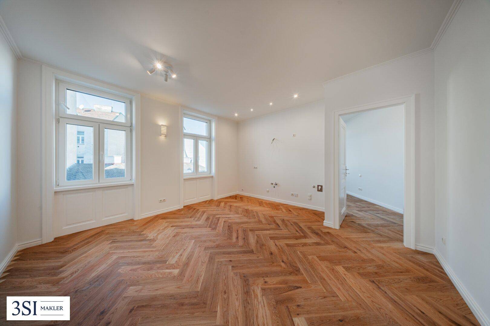 Geräumiges Zimmer mit Fischgrätparkett, zwei Fenstern und Zugang zu einem weiteren Raum.