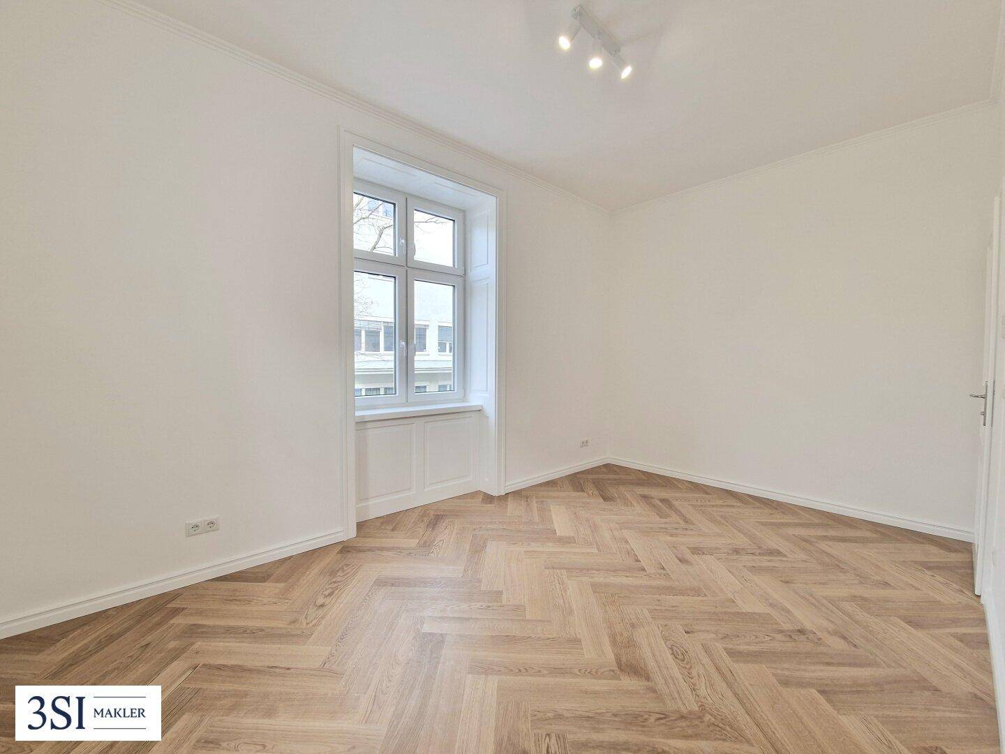 Helles Zimmer mit Fischgrätparkett und einem großen Fenster, ideal als Schlaf- oder Arbeitszimmer.