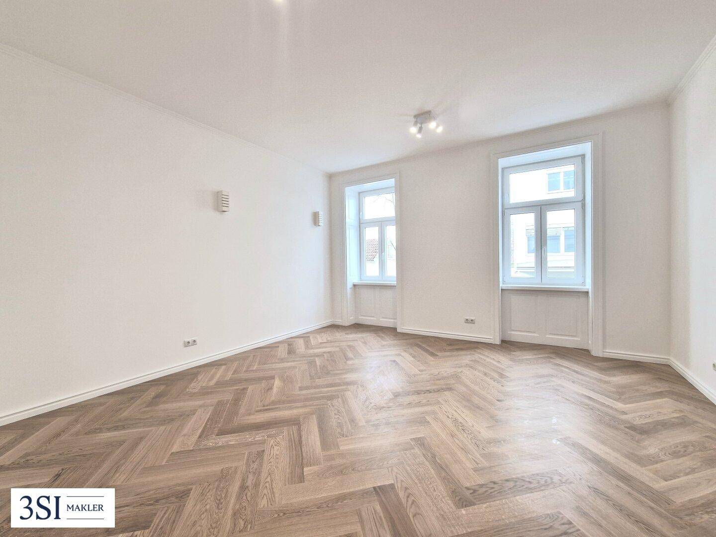 Großes, helles Zimmer mit Fischgrätparkett und zwei hohen Fenstern, ideal für Wohn- oder Essbereich.