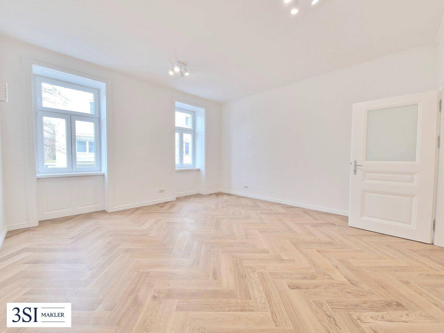 Helles Zimmer mit Fischgrätparkett, zwei Fenstern und einer modernen Tür mit Glaseinsatz.