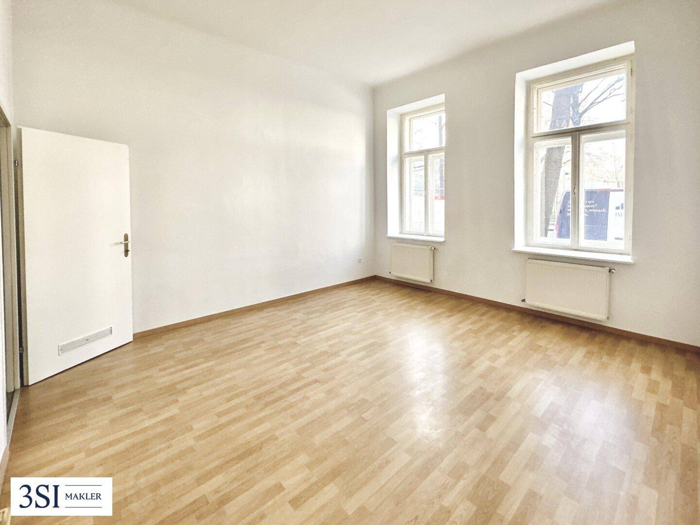Heller, leerer Raum mit Parkettboden, weißen Wänden und zwei Fenstern mit Heizkörpern.