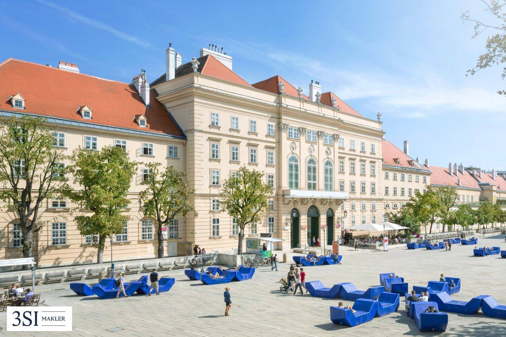 Prächtiges historisches Gebäude an einem weitläufigen Platz mit modernen blauen Sitzgelegenheiten.