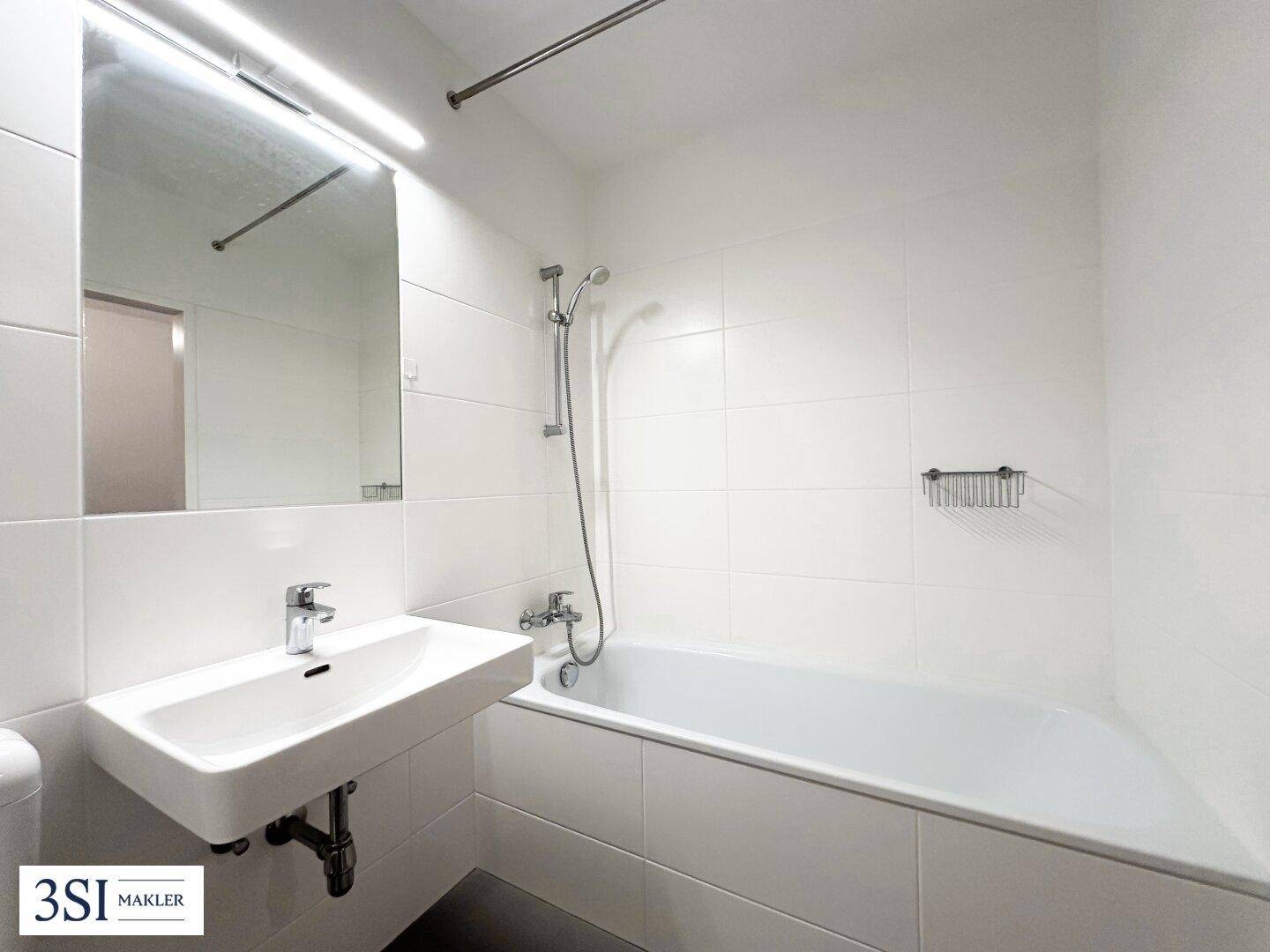 Helles Badezimmer mit weißen Fliesen, Badewanne mit Duschfunktion und modernem Waschbecken.