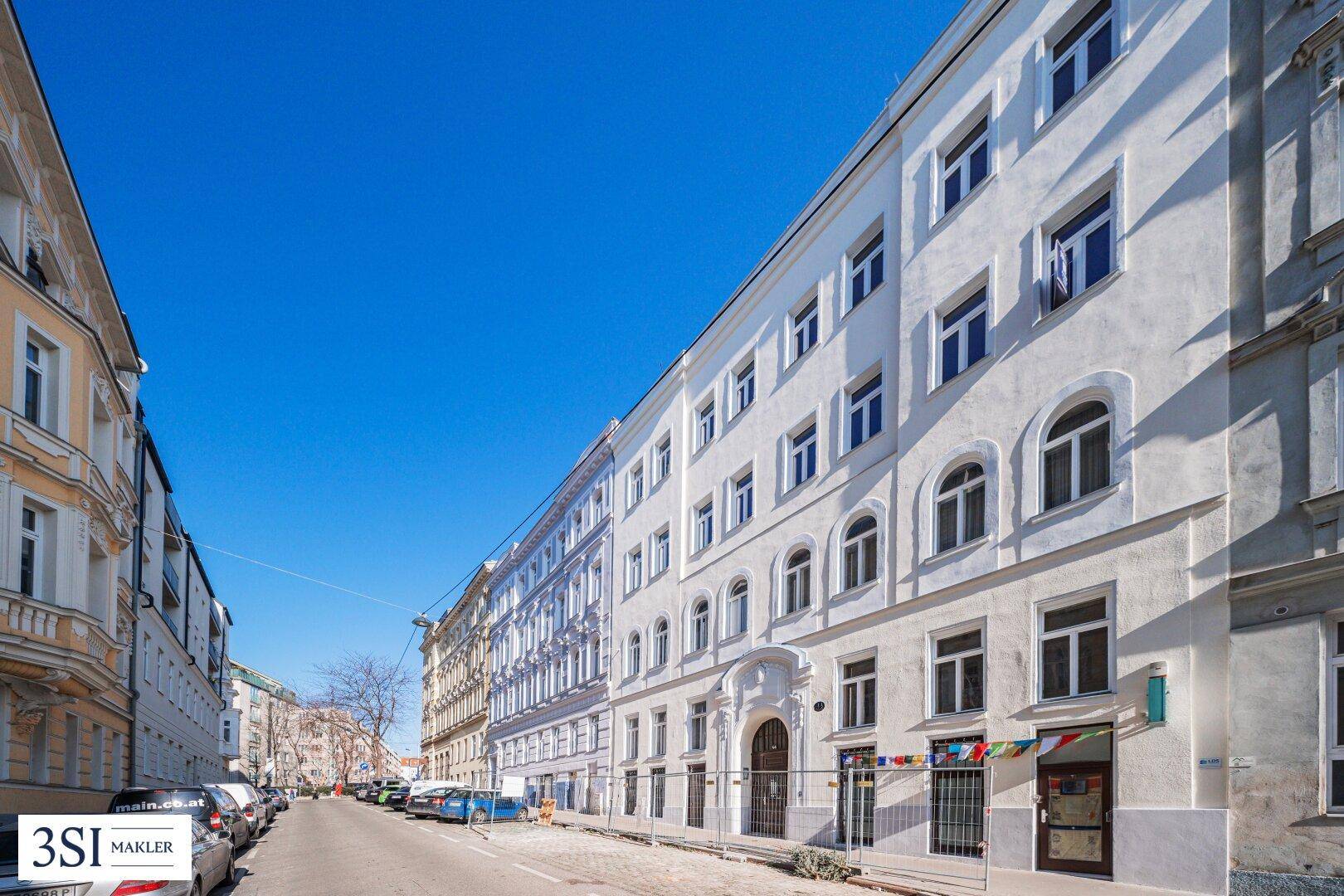 Lange weiße Gebäudefassade an einer städtischen Straße mit parkenden Autos unter strahlend blauem Himmel.