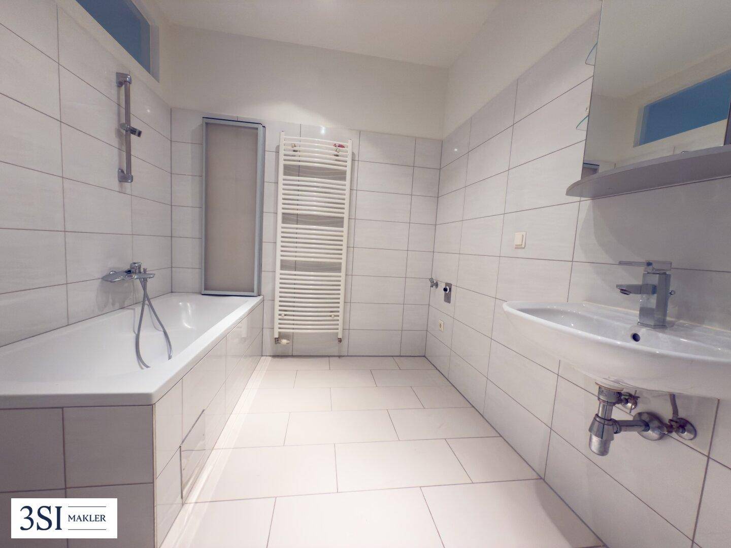 Helles Badezimmer mit weißen Fliesen, einer Badewanne und einem praktischen beheizten Handtuchhalter.