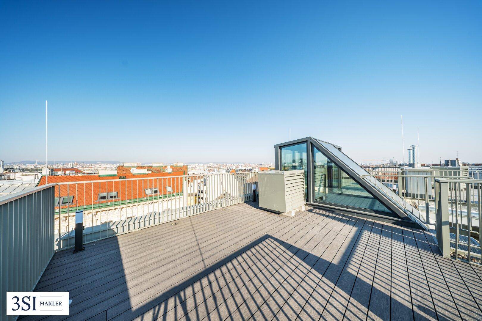 Geräumige Dachterrasse mit Holzboden, Glasaufbau und weitem Stadtblick unter blauem Himmel.