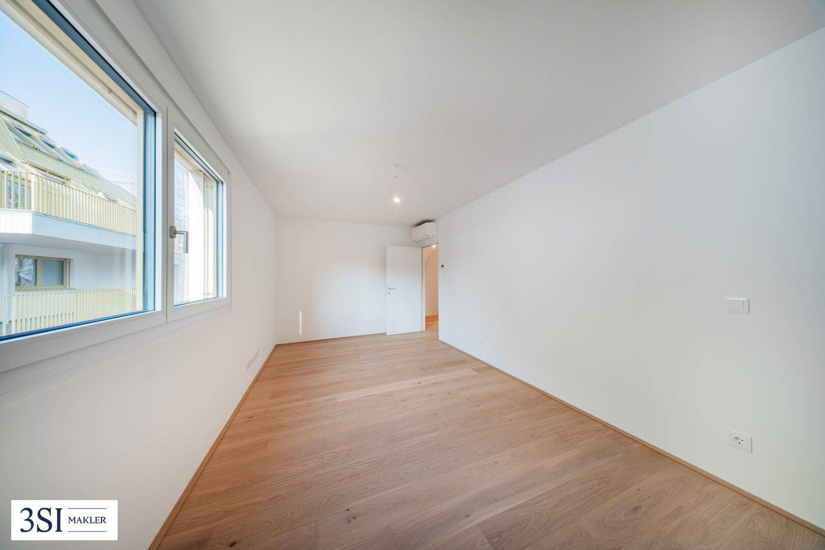 Helles, leeres Zimmer mit Holzboden, weißen Wänden und großen Fenstern.
