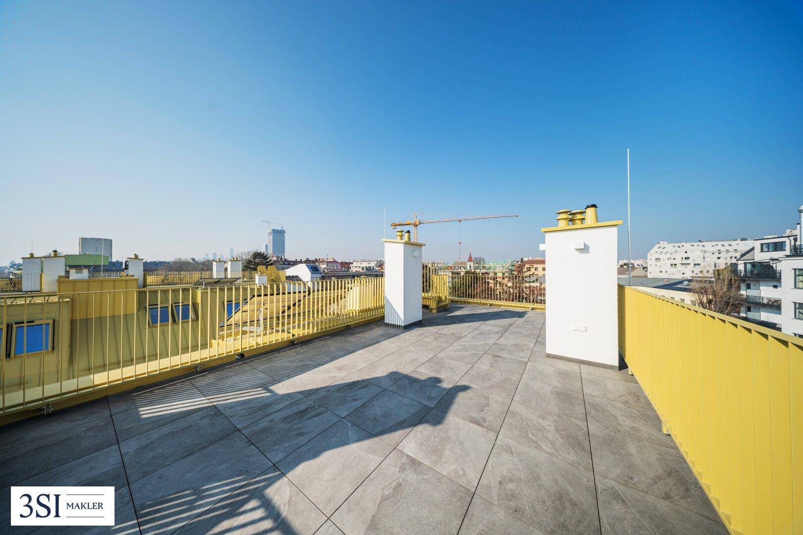 Weitläufige Dachterrasse mit Fliesenboden und gelbem Geländer unter blauem Himmel.
